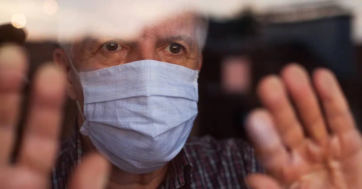 An old white man wearing a medical mask looking out on the window