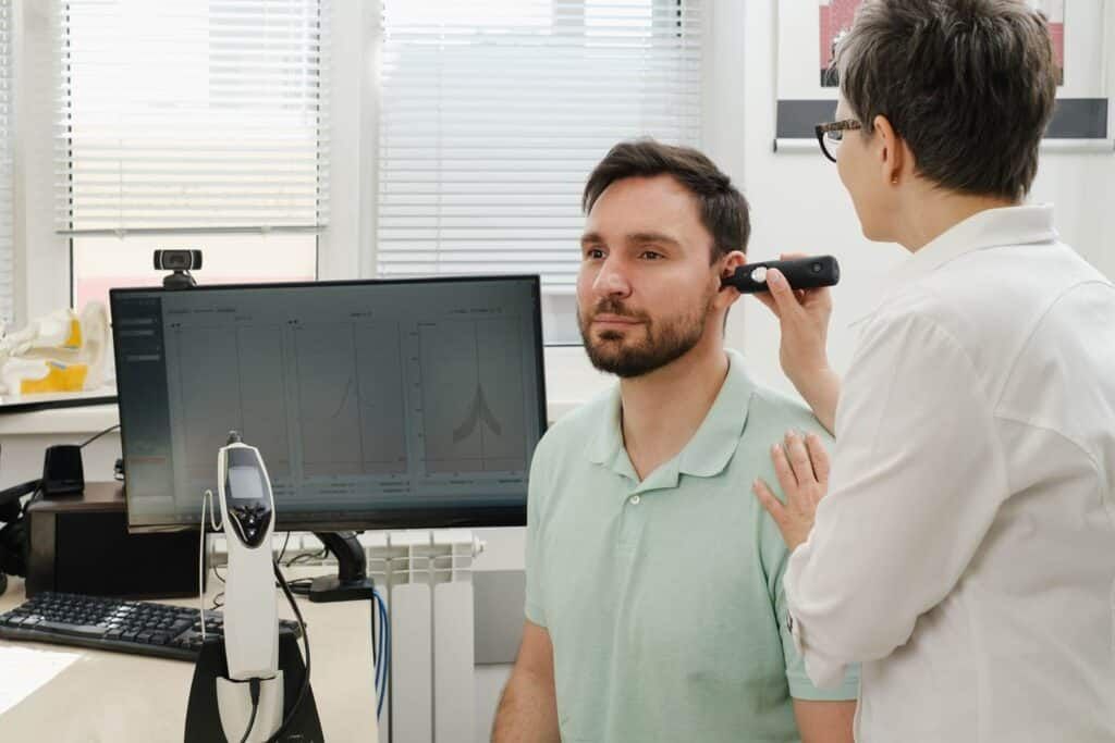 Male patient receiving an ear examination to check on VA disability for tinnitus symptoms, as a healthcare professional uses an otoscope in a clinical setting with diagnostic data displayed on a computer screen in the background.